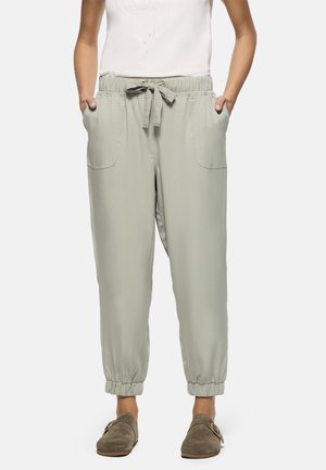 Pantalones casuales gris claro con puños elásticos y cintura con cordón, usados con zapatos tipo mocasín taupe; manos descansando en los bolsillos delanteros.