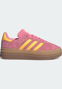 Różowe zamszowe sneakersy z żółtymi akcentami, z trzema paskami i teksturowanym różowym uchwytem. Gruba przezroczysta podeszwa z brandingiem.