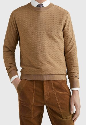 Mann in beigem gemustertem Pullover, weißem Hemd mit Kragen und gestreifter Krawatte sowie braunen Cordhosen, mit den Händen in den Taschen.