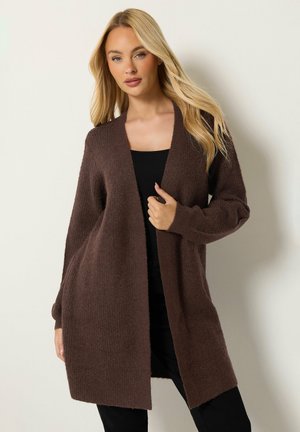 Blonde vrouw die een lange, bruine open cardigan draagt over een zwart topje en een zwarte broek, staand tegen een effen lichte achtergrond.