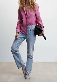 Femme portant une chemise rose à manches longues, un jean bleu clair et des ballerines argentées, tenant un sac à main noir sur un fond uni.