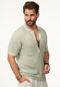 Hellgrünes, kurzärmliges, gestricktes Strickjäckchen mit offenem Netzdesign, Knopfverschluss und gerippten Akzenten an Kragen und Bündchen.