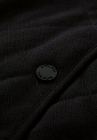 Czarna pikowana tkanina z wyraźnym okrągłym czarnym guzikiem z wytłoczonym napisem "Tom Tailor" oraz teksturowanym ukośnym przeszyciem.