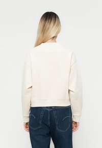 Donna con lunghi capelli biondi che indossa una semplice felpa bianca sporca e jeans blu scuro, girata di spalle su uno sfondo bianco.