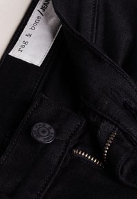 Jeans de mezclilla negra con una etiqueta de marca visible, cremallera de metal y botón de tela. La textura es suave con detalles de costura sutiles.