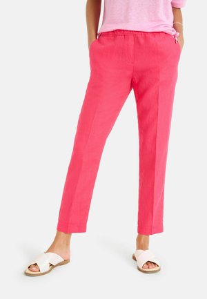 Frau trägt knallpinke, gerade geschnittene Hose und weiße flache Sandalen mit überkreuzten Riemen, steht mit Händen in den Taschen vor schlichtem Hintergrund.