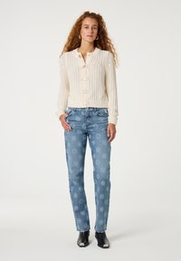 Crème gebreide cardigan met knoopsluiting, gecombineerd met lichtblauwe floral-geprinte jeans en zwarte enkellaarsjes met metallic accenten.