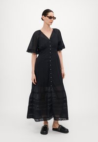 Abito lungo nero con bottoni, maniche a campana e orlo a balze. Realizzato in tessuto leggero con dettagli in pizzo. Abbinato a semplici sandali neri.