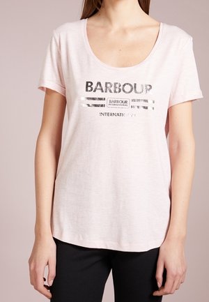 Kobieta w jasno różowej koszulce Barbour International z czarnym napisem i paskami, w połączeniu z czarnymi spodniami, stoi na jednolitym tle.