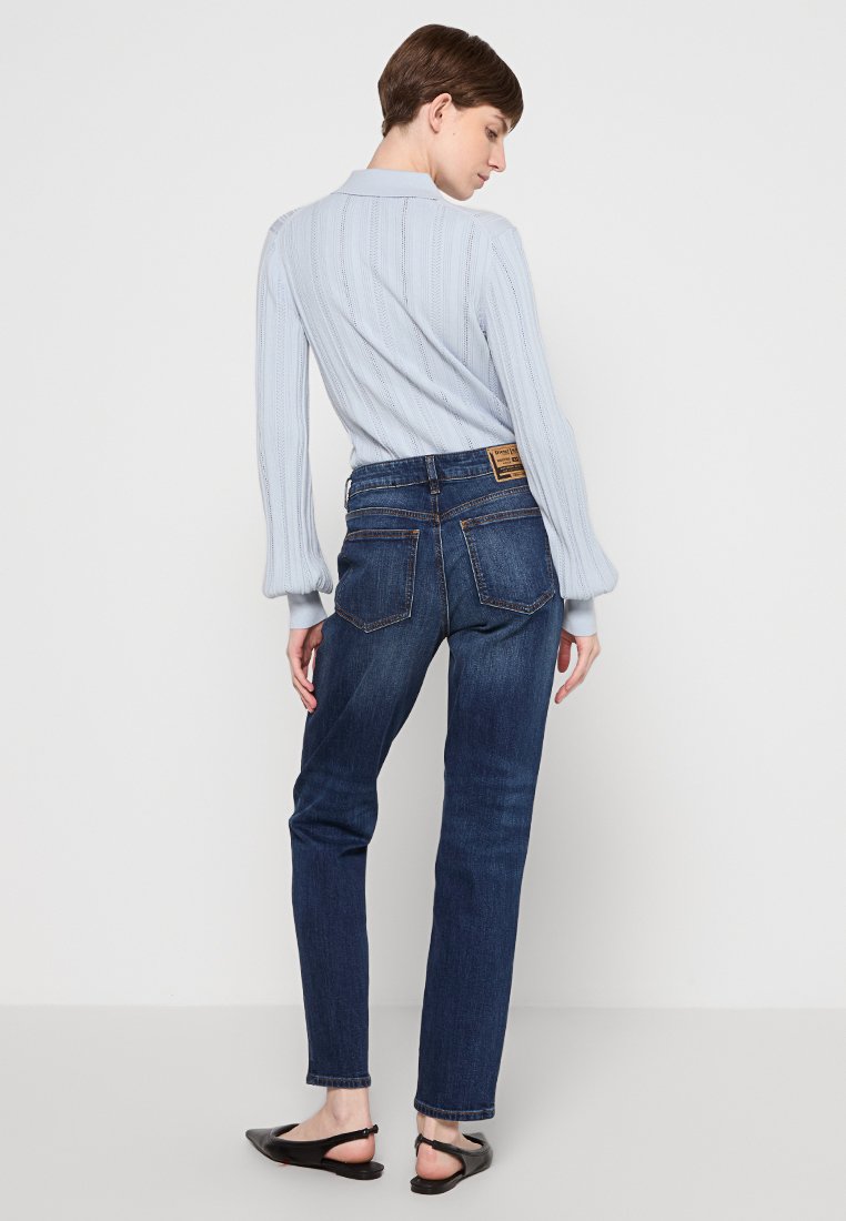 Donna in piedi di spalle, che indossa un maglione lavorato a maglia di colore blu chiaro, jeans dritti blu scuro e ballerine nere.