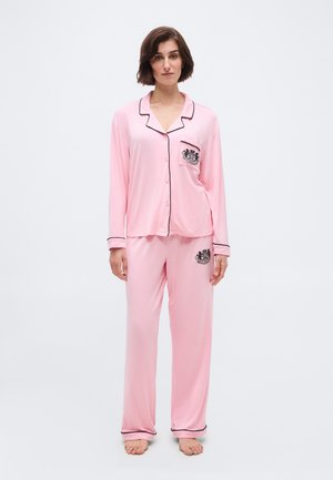 Jalal seisev naine, kes kannab paljajalu valgelt roosat pika varrukaga nööbitavat pidžaamat mustade servadega ja tikandiga embleemidega taskul ning pükstel.