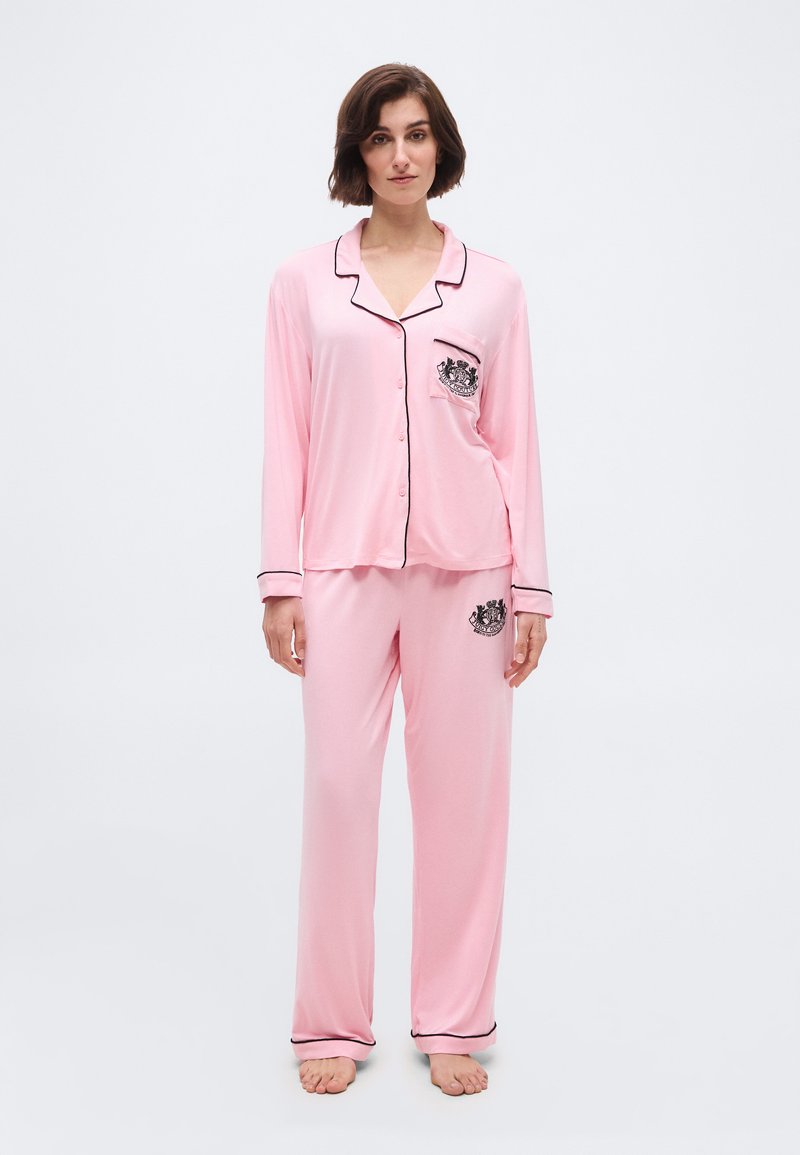 Γυναίκα που στέκεται ξυπόλυτη, φορώντας ροζ ανοιχτό ροζ πιτζάμες με μακριά μανίκια και κουμπιά, με μαύρη διακόσμηση και κεντημένα σήματα στην τσέπη και το παντελόνι.