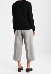 Osoba stoi tyłem, ubrana w czarną bluzkę z długim rękawem, szare szerokie spodnie o długości do kostek i czarne buty oxford na białym tle.