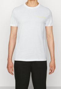T-shirt bianco a maniche corte in cotone, con scollo rotondo e logo giallo "HILFIGER" sul lato sinistro del petto. Texture liscia, vestibilità regolare.