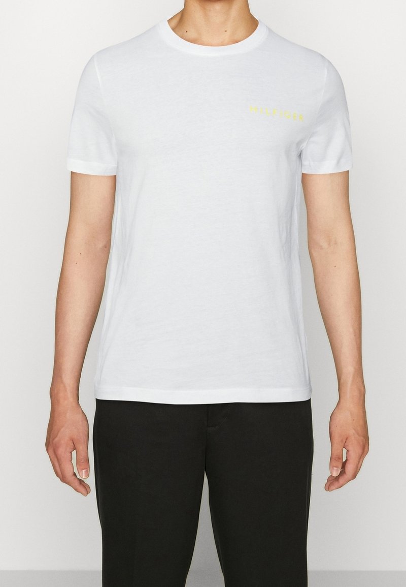 T-shirt bianco a maniche corte in cotone, con scollo rotondo e logo giallo "HILFIGER" sul lato sinistro del petto. Texture liscia, vestibilità regolare.