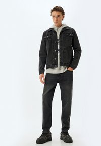 Chaqueta de denim negra con bolsillos frontales, combinada con una sudadera gris con un estampado gráfico, pantalones de denim negros y zapatillas oscuras.