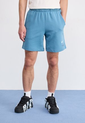 Mężczyzna ubrany w niebieskie sportowe szorty Nike, czarno-białe buty sportowe Nike, stoi na niebieskiej podłodze z rękami w kieszeniach.