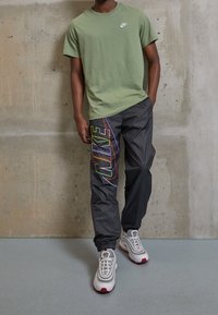 Man som bär olivgrön Nike-t-shirt, svarta Nike-byxor med flerdelad logga och vita Nike-sneakers, stående mot en betongvägg.