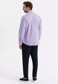 Camisa de manga larga lila claro con puños abotonados, corte recto, pliegue en la parte trasera central, combinada con pantalones oscuros y zapatos negros sin cordones.