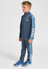 Blauwe trainingsbroek met een half-ritstop en bijpassende broek met lichtblauwe accenten en witte chevronpatronen op de mouwen en zijkanten.