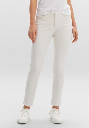 Jean slim beige clair en coton mélangé, avec une fermeture à bouton, cinq poches, et des baskets blanches. Texture lisse.