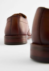 Chaussures en cuir marron avec une texture lisse et de subtils détails de couture, dotées d'une semelle en bois plate et d'un bout arrondi.