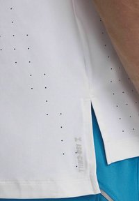 Camiseta deportiva blanca con pequeños puntos azules, con costuras laterales ventiladas y una etiqueta "ISO-CHILL" cerca del dobladillo. Textura de tela suave.