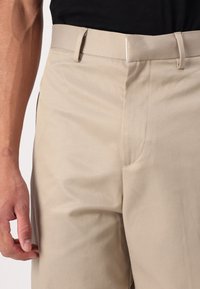 Pantalon beige pour homme avec passants de ceinture, fermeture avant dissimulée, porté avec un haut noir, présenté de la taille à mi-cuisse.