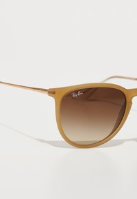 Ochelari de soare Ray-Ban cu o ramă mată de culoare miere, lentile maro degradé și accente metalice. Au o formă rotundă și brațe subțiri.