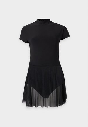 Vrouw draagt een zwarte jurk met korte mouwen en hoge hals met een doorzichtige geplooide rok en zwarte sandalen met hoge hakken en bandjes, staat rechtop.