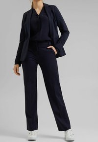 Námořnický modrý sako na míru přes odpovídající top a kalhoty. Materiál vypadá hladce. Bílé tenisky dokonale doplňují outfit.