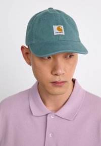 Teal bomullslue med buet skjerm, som har et rektangulært hvitt merke med en gul logo. Båret over en lys lilla polo-skjorte.