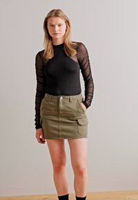 Svart langermet bodysuit med gjennomsiktige ermer, kombinert med en olivengrønn høyt liv-skjørt, som har sidelommer og knappelukking.
