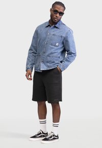 Veste en denim avec une poche poitrine, deux poches avant et fermeture à boutons ; associée à un short noir et des baskets noires avec des accents blancs.