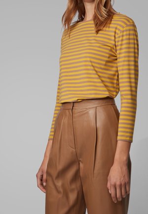 Kobieta w żółto-brązowej pasiastym długim rękawie, wpuszczona w brązowe, skórzane spodnie z wysokim stanem, stojąca na jednolitym szarym tle.