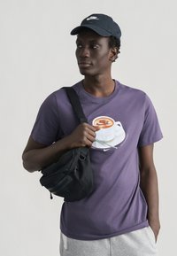 Νεαρός άνδρας φοράει μαύρο καπέλο Nike, μωβ μπλουζάκι Nike με γραφικό κούπα καφέ, γκρι παντελόνι και μία μαύρη τσάντα ώμου.