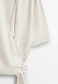 Top avvolgente bianco realizzato in tessuto liscio, con un accentuato nodo laterale e maniche a metà con una leggera caduta.
