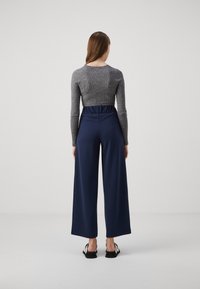 Femme aux longs cheveux bruns portant un haut gris côtelé à manches longues, un pantalon large bleu marine et des chaussures plates noires et blanches, de dos.
