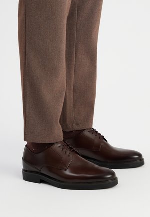 Zapatos de vestir de cuero marrón con un acabado suave, cierre con cordones y suela de goma oscura, combinados con pantalones de vestir marrones y calcetines oscuros con un patrón.
