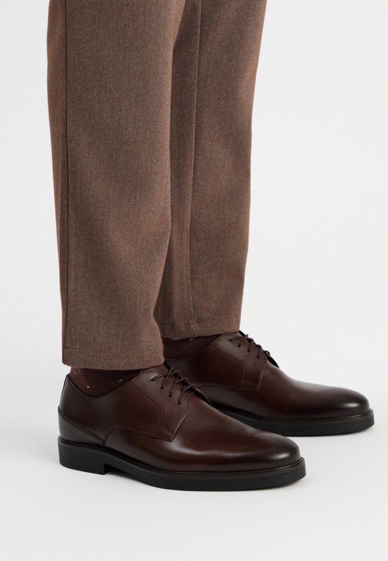 Scarpe eleganti in pelle marrone con una finitura liscia, chiusura con lacci e suola in gomma scura, abbinate a pantaloni eleganti marroni e calzini scuri a fantasia.