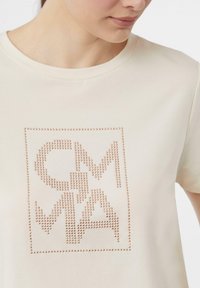Cream-colored t-shirt featuring a square studded design with "CMMA" letters in a textured, metallic finish. Soft fabric and crew neck.