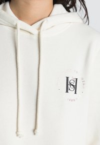 Weißer Hoodie mit Kordelzügen, ausgestattet mit einem runden Logo in schwarzer und roségoldener Schrift. Weicher Stoff mit lockerem Schnitt und Kängurutasche.