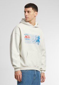 Hanorac cu glugă de culoare gri deschis, cu un buzunar frontal, având un design grafic "ACL Athletics" în culorile albastru și roșu și manșete ribbate.