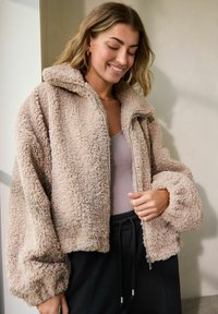 Lysbrun fleece zip-up jakke med høj krave, oversized pasform og elastiske manchetter. Båret over en grå top og mørke bukser.
