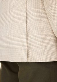 Beżowy marynarka w kratę zachodząca na ciemnozielone spodnie, ukazująca fakturę tkaniny i kontrast kolorów na krawędziach ubrań.