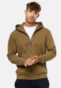 Olivengrøn zip-up hoodie med en tekstureret bomuldsblanding, ribbede manchetter og elastisk kant. Har en kængurulomme og et logo på brystet.