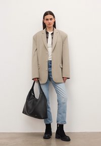 Naine, kes kannab üle suuruse beeži bleiserit, valget särki, ruudulist sisekihist, helesinisi teksaseid ja musti saapaid, hoiab suurt musta käekotti ning seisab ühetaolise taustaga.