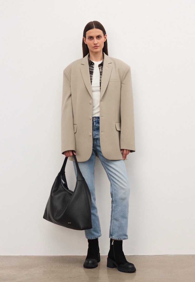 Naine, kes kannab üle suuruse beeži bleiserit, valget särki, ruudulist sisekihist, helesinisi teksaseid ja musti saapaid, hoiab suurt musta käekotti ning seisab ühetaolise taustaga.