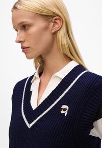 Navy knitted vest with a v-neckline, white trim, and a small embroidered logo on the left side. Worn over a white collared shirt.