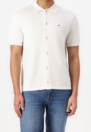 Homme portant un polo blanc à manches courtes avec boutons, avec un petit logo sur le côté gauche de la poitrine et un jean bleu.